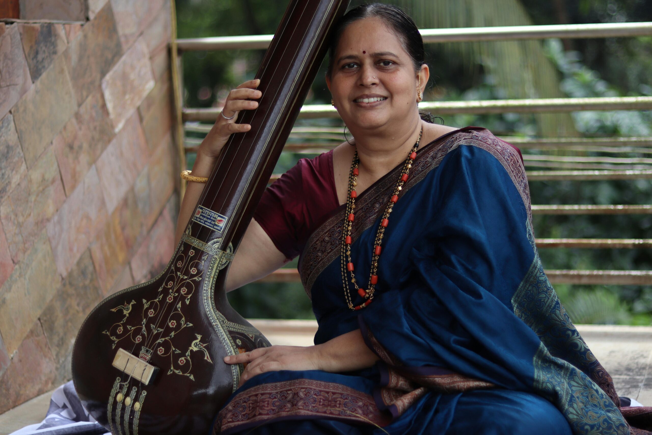 Smiling woman in traditional Indian attire holding a tanpura outdoors.