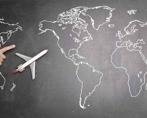 Top view of miniature airplane placed on over gray world map with crop hand of anonymous person indicating direction representing travel concept Abroad